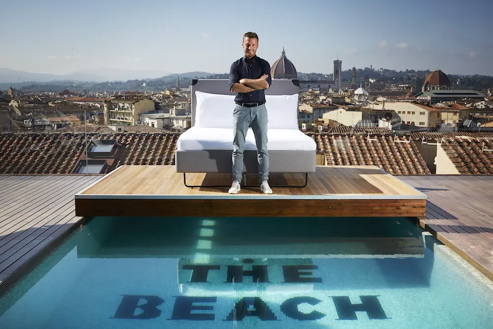 Charlie MacGregor, chief executive officer and founder of The Student Hotel (TSH) at the opening of his first Italian hybrid hotel, TSH Florence Lavagnini. Charlie is pictured standing in front of a bed on a platform above the rooftop pool in front of the Duomo in the Tuscan capital, Florence.